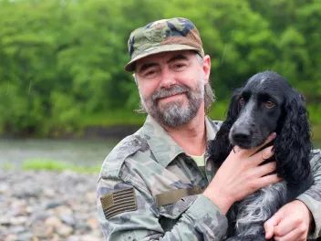 a man in army fatigues holds a dog while outside by a river bank